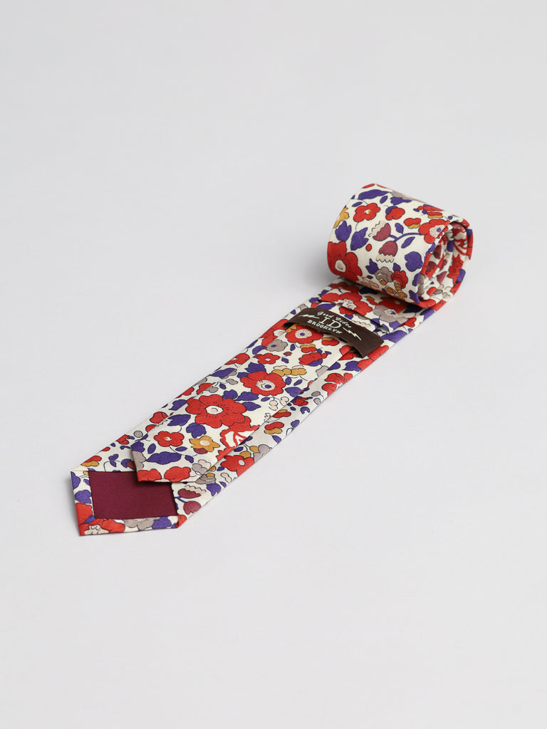 The Liberty of London Tana Lawn™ Betsy Tie by ID Menswear, handmade in Brooklyn from soft Tana Lawn cotton, features a colorful floral pattern with red, purple, and yellow flowers on white, displayed rolled on a plain white surface.