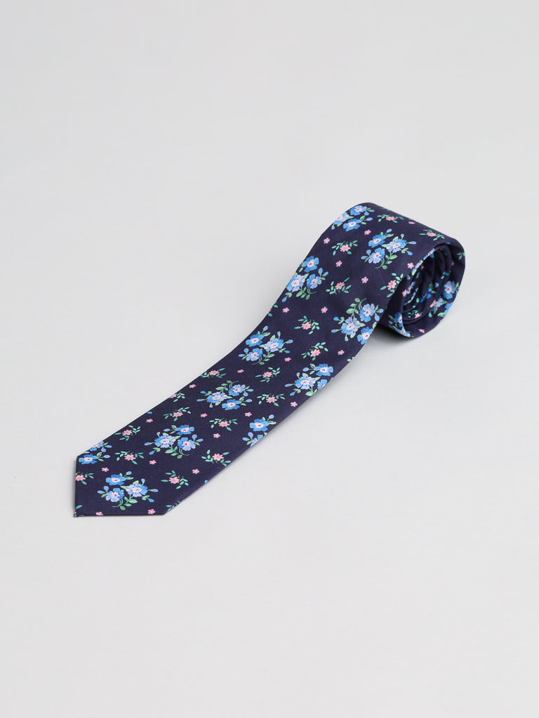 A rolled-up Liberty of London Tana Lawn™ Magdalena Tie by ID Menswear, handmade in Brooklyn from cotton, with a blue and white floral pattern and green leaves, displayed on a light gray background.