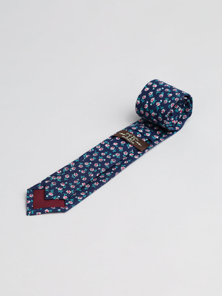 The White Bloom Cotton Tie by ID Menswear is a handmade navy blue cotton tie with red, white, and light blue floral patterns, shown rolled up against a plain white background.