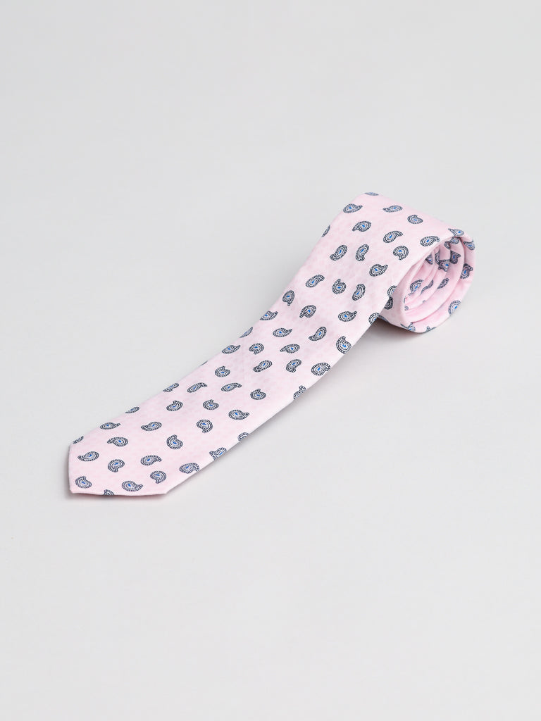 The Bombay Cotton Tie by ID Menswear, handmade in Brooklyn, is a pale pink cotton tie with a repeating blue paisley pattern, shown rolled-up on a plain light gray background.