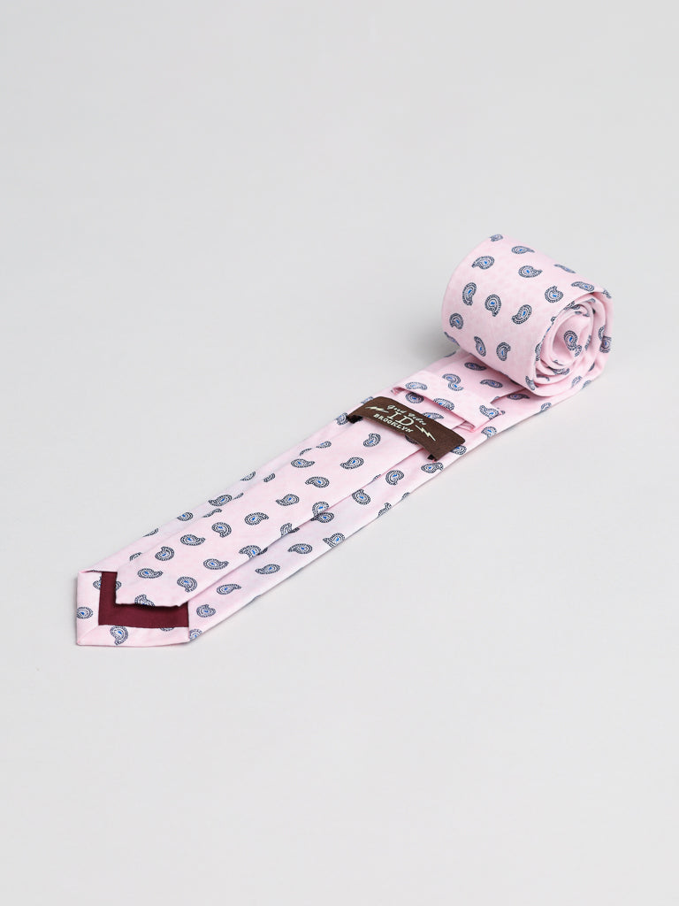 The Bombay Cotton Tie by ID Menswear is a light pink cotton tie with a blue paisley pattern, partially unrolled on a plain light gray background. Handmade in Brooklyn, it’s detailed with a brown label—ideal for business formal occasions.