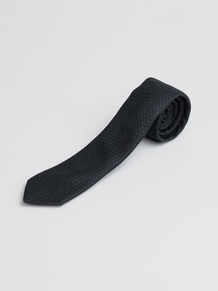 The Chicago Cotton Tie by ID Menswear, a handmade black textured cotton tie crafted in Brooklyn, USA, is shown rolled up on a light gray background—a timeless business formal accessory.