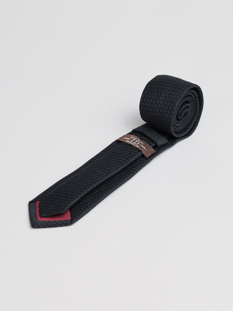 The ID Menswear Chicago Cotton Tie, a textured black handmade accessory with a rolled end and red-accented tip, is displayed on a light background. A brown label adds detail, making it perfect for business formal looks with a Brooklyn USA vibe.