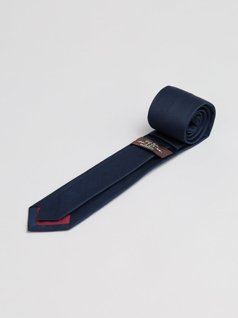 The ID Menswear Midnight Cotton Tie, handmade in Brooklyn in dark navy blue cotton, is rolled and laid flat on a light gray surface with a brown tag near the tip, which reveals a subtle dark red accent underneath.