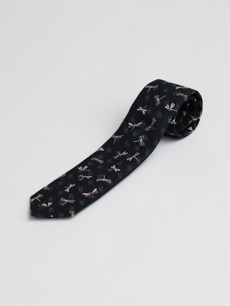 The Japanese Printed Dragonfly Tie by ID Menswear, handmade in Brooklyn from 100% cotton, is shown unrolled on a plain light gray background and features a black base with a colorful dragonfly print.