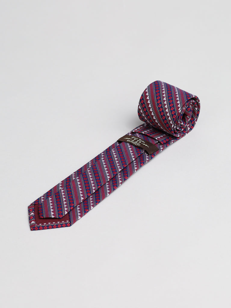 The Karuna Cotton Tie by ID Menswear, featuring a red, blue, and white geometric pattern, is handmade in Brooklyn and shown rolled up on a plain white background.