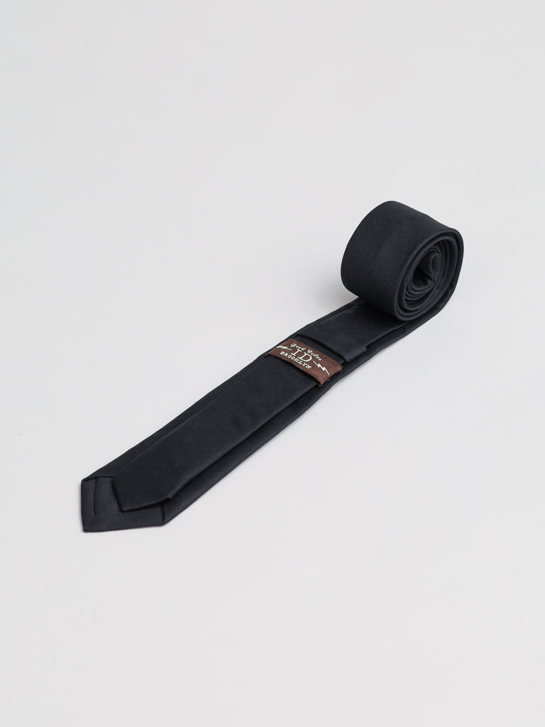 The Super Skinny Cotton Tie by ID Menswear, handmade in Brooklyn from black cotton with a brown and gold label, is displayed on a plain white background—ideal for business formal occasions.