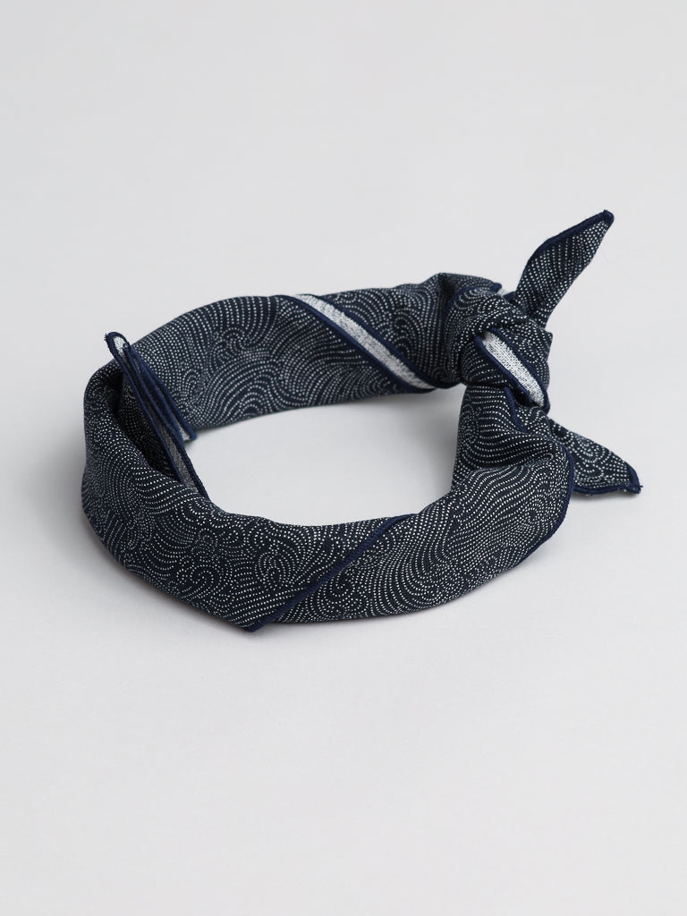 The Waves and Water Bandana from ID Menswear, featuring an intricate white pattern on navy 100% cotton, is rolled and loosely knotted on a plain light gray background.