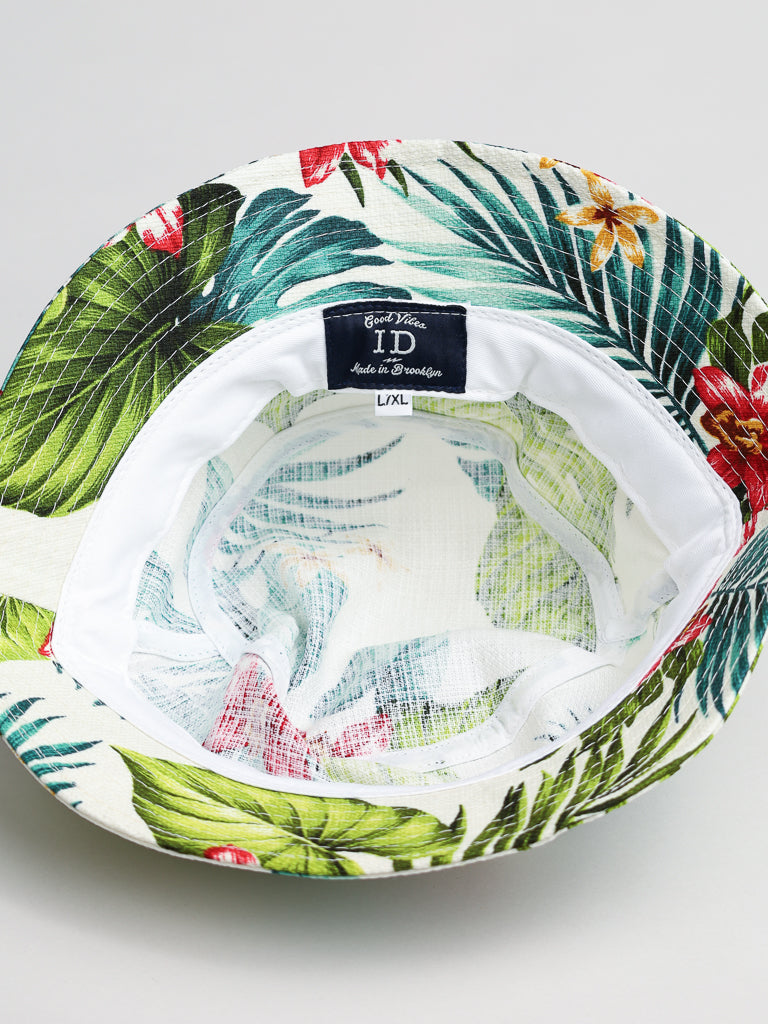 The ID Hawaii Bucket Hat by ID Menswear is crafted from Japanese cotton and features a tropical pattern with green leaves and red and yellow flowers. Made in Brooklyn, it includes an upside-down view revealing a size tag L/XL inside.
