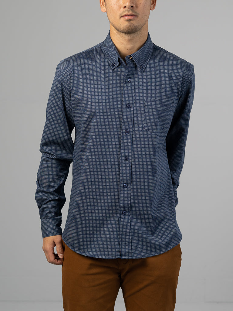 Against a gray backdrop, an individual in brown pants dons an ID Menswear Leo Stretch Button Down Shirt, known for its high-quality finish and subtle blue checkered pattern. The shirt has long sleeves and offers comfortable stretch, as the person looks slightly to the side.