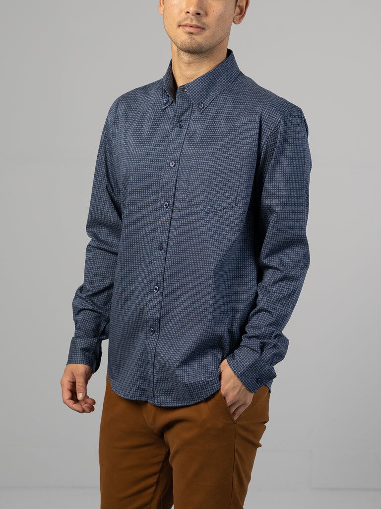 Wearing a Leo Stretch Button Down Shirt by ID Menswear, featuring a subtle checkered design, long sleeves, and collar for style and comfort, paired with brown pants against a gray background.
