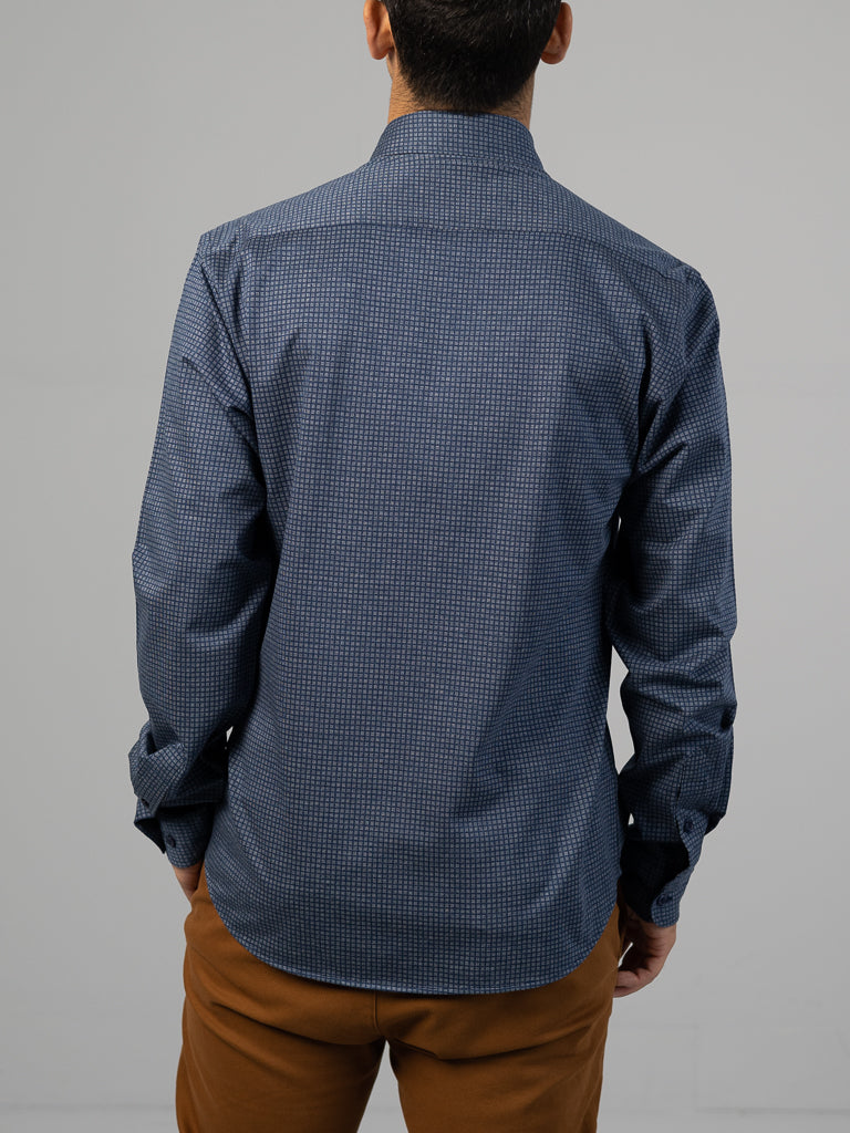 A person faces away from the camera wearing an ID Menswear Leo Stretch Button Down Shirt in dark blue, known for its comfortable stretch and high-quality finish, alongside brown pants. The setting is a simple light-colored wall.