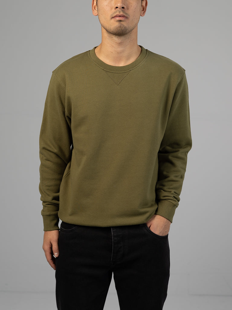 A person in an olive green Everyday French Cotton Terry Crewneck Sweatshirt by ID Menswear and black pants stands against a plain gray background, one hand casually in their pocket, with their face partially obscured.