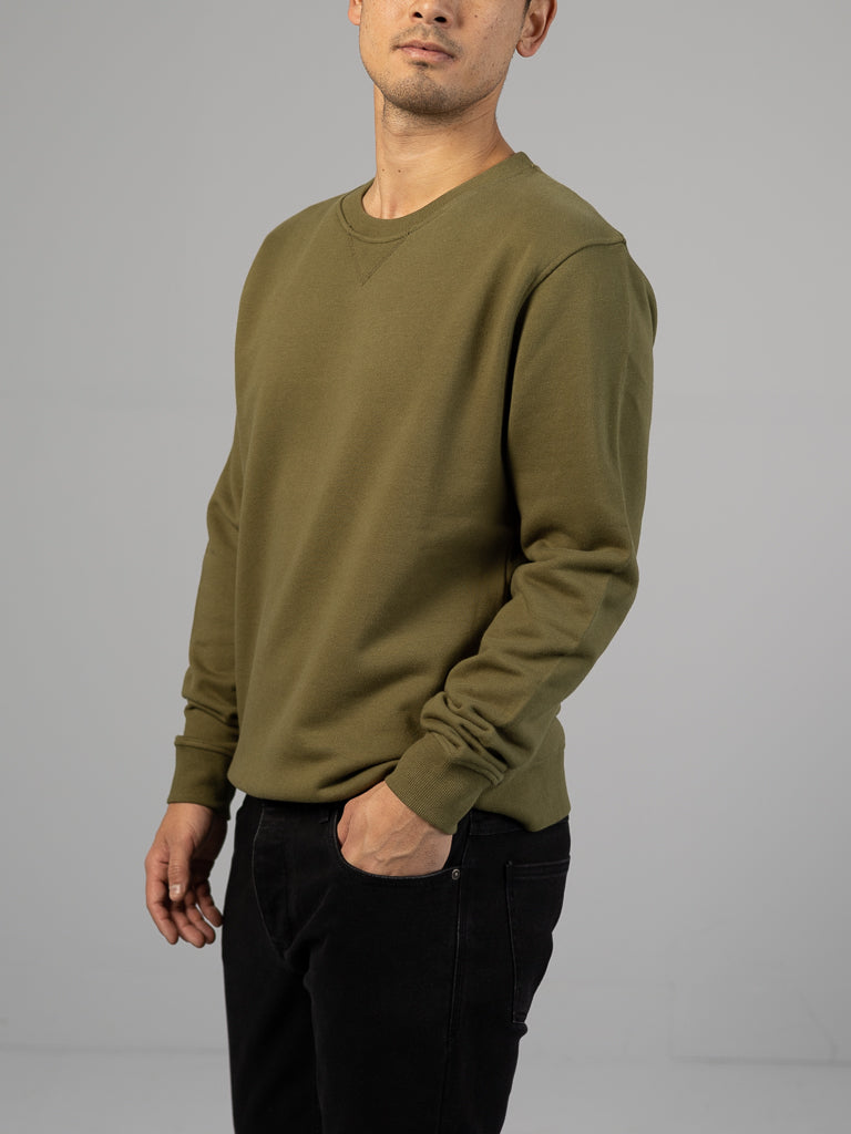 Against a gray backdrop, someone models an ID Menswear outfit: the Everyday French Cotton Terry Crewneck Sweatshirt in olive green paired with black pants, one hand in their pocket and the other at their side, while minimal facial features are visible.