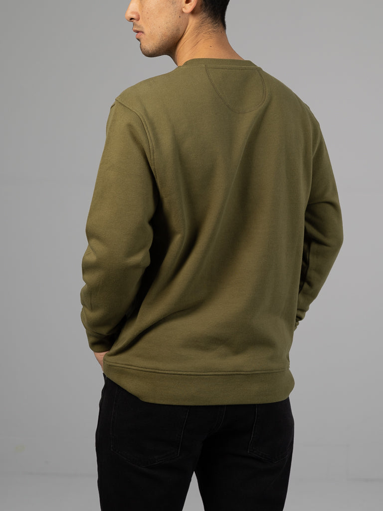 A person in a green Everyday French Cotton Terry Crewneck Sweatshirt from ID Menswear and black pants stands against a plain gray background, viewed from behind, slightly turned to the left, with hands at their sides.