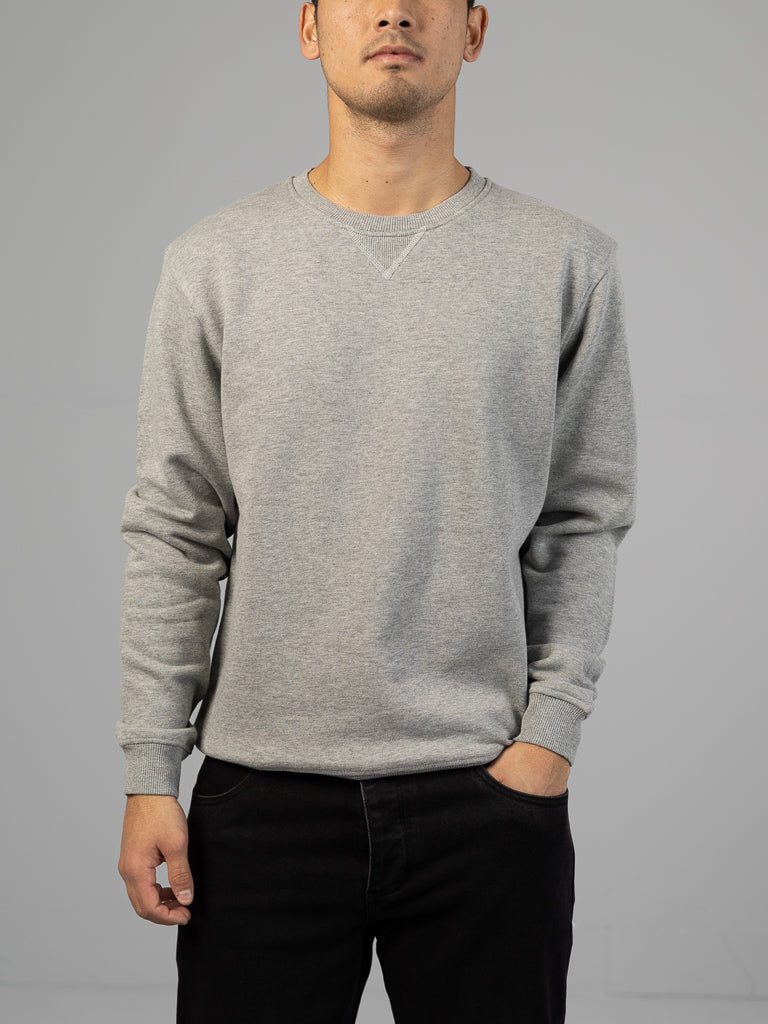 A person in a plain, light gray Everyday French Cotton Terry Crewneck Sweatshirt by ID Menswear, crafted from cozy French cotton terry, stands with one hand in a pocket against a neutral backdrop. The sweatshirt boasts subtle stitch detailing at the collar.