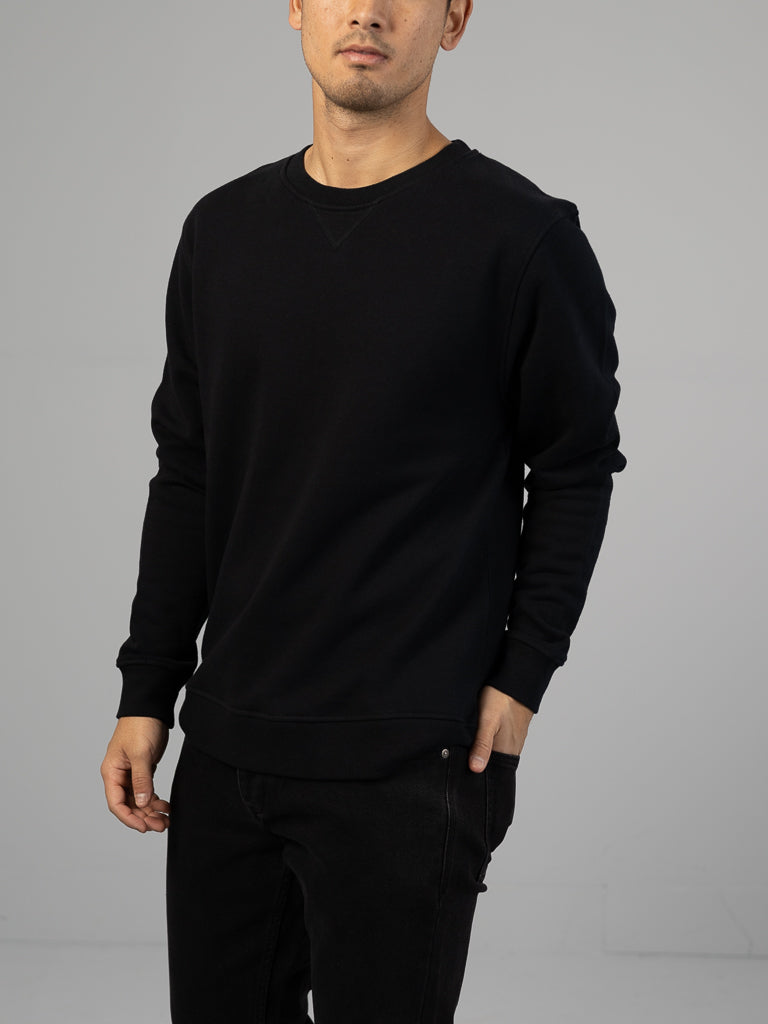 A person dressed entirely in black wears an Everyday French Cotton Terry Crewneck Sweatshirt by ID Menswear, posing with one hand in their pocket against a plain gray background, their face obscured.