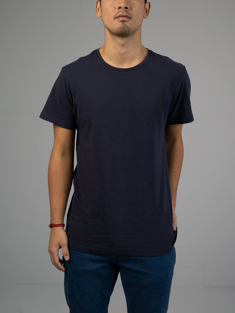 A person stands against a neutral background wearing the Form Crew Neck Tee from ID Menswear in dark blue, paired with blue jeans. The outfits classic and timeless style speaks volumes, even without the persons head in the frame.