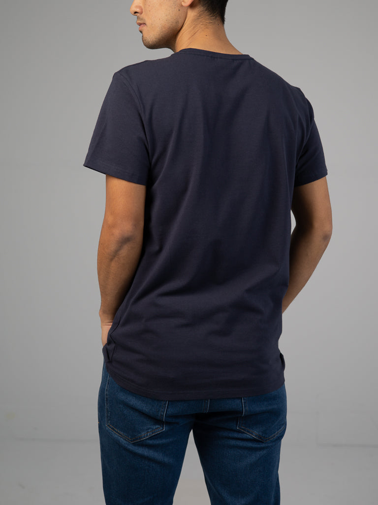 A person stands with their back to the camera, wearing a dark blue Form Crew Neck Tee by ID Menswear and jeans. The classic design complements the simple style against the plain light gray background.