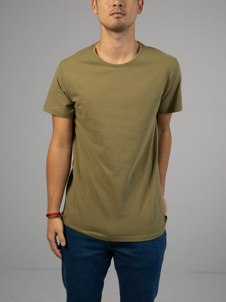 A person stands against a neutral background wearing an ID Menswear Form Crew Neck Tee in olive green and blue jeans, with a red bracelet on their wrist. Their relaxed stance emphasizes the comfortable fabric of the tee.