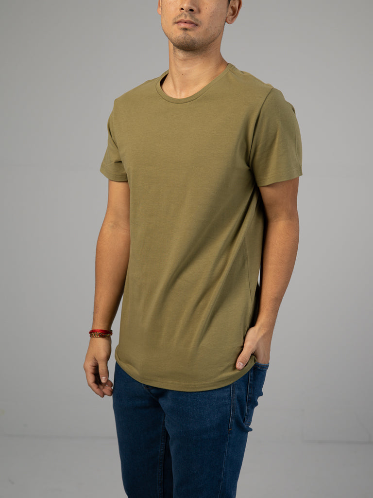A person in an ID Menswear Form Crew Neck Tee in olive green and blue jeans casually stands with a hand in a pocket against a plain light gray background, obscuring their face.