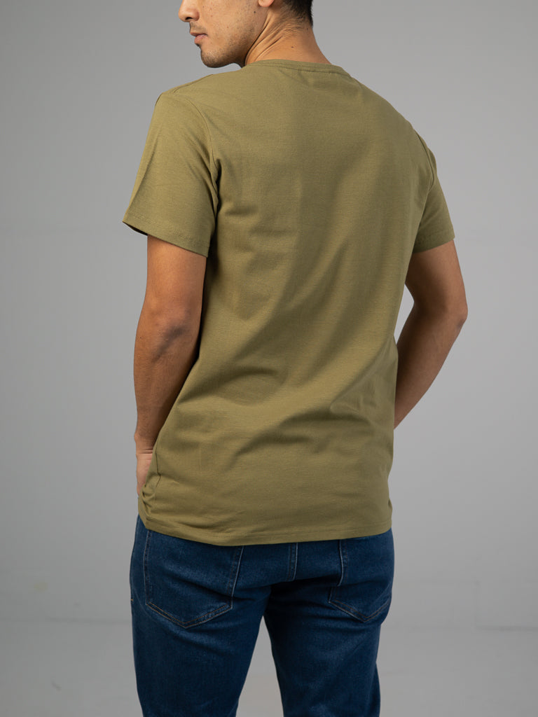 A person wearing the ID Menswear Form Crew Neck Tee in khaki paired with blue jeans stands with their back to the camera, hand in pocket, highlighting its form-fitting style against a plain gray background.