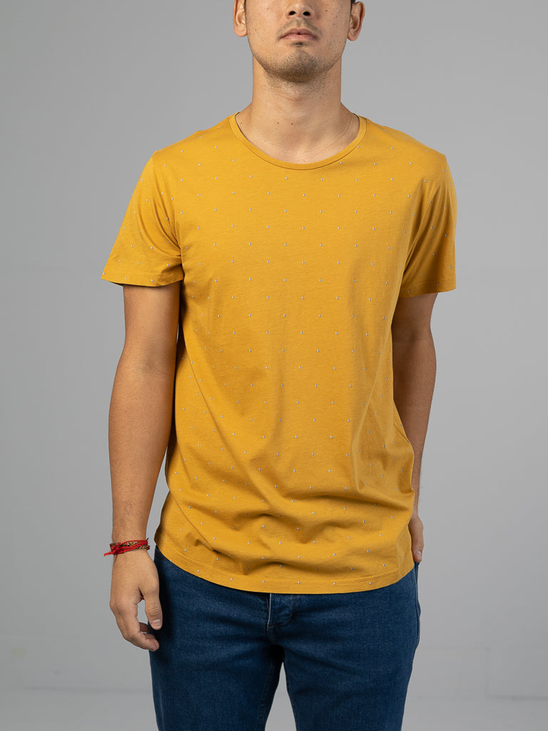 Against a gray background, someone sports the Heritage Printed Organic Cotton Tee by ID Menswear in classic fit yellow with small dots and blue jeans. Hands in pockets, a red bracelet adorns their wrist, though their face is obscured.