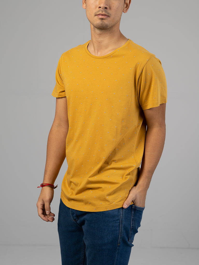 A person in a mustard yellow Heritage Printed Organic Cotton Tee by ID Menswear, featuring small white dots, pairs it with blue jeans. They stand against a plain gray background, left hand in pocket and a red bracelet on their right wrist.