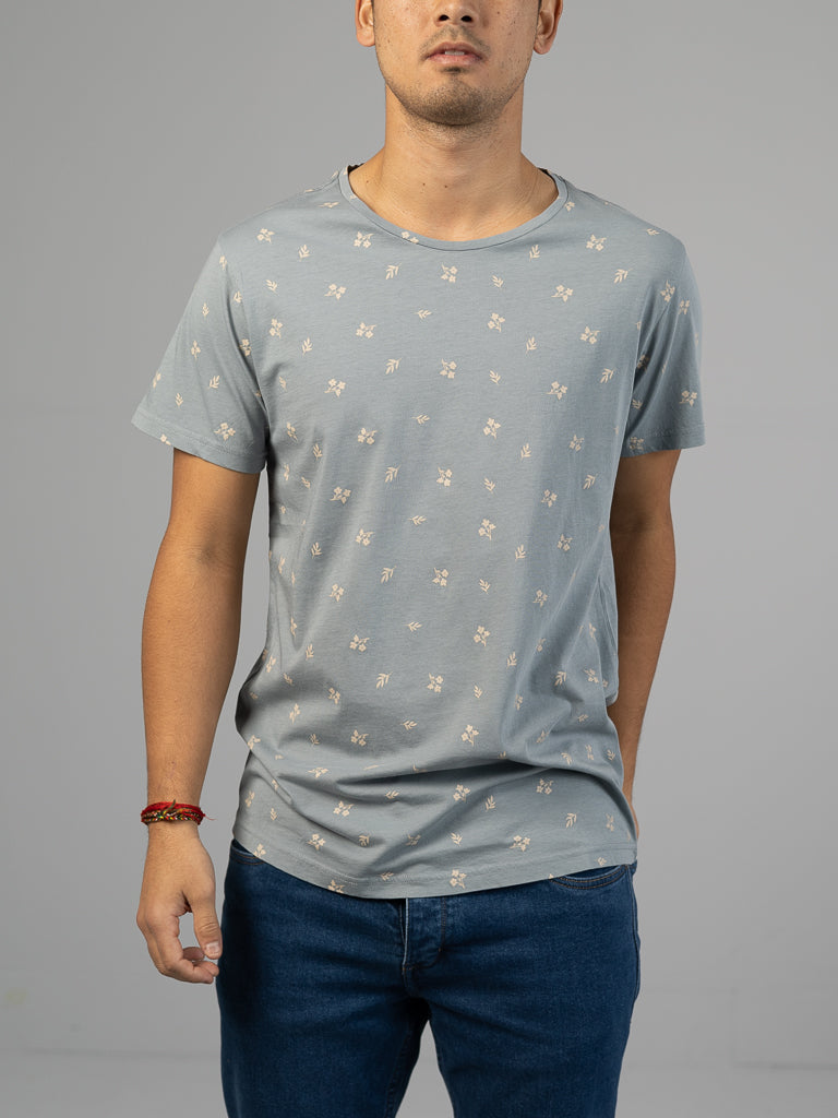 Against a neutral background, a person models the ID Menswears Heritage Printed Organic Cotton Tee in light blue with a floral pattern, paired with dark blue jeans. A red beaded bracelet adorns their wrist, while their face is not shown.