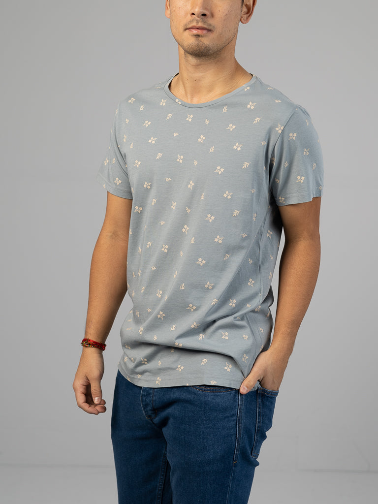 A person stands against a grey backdrop wearing an ID Menswear Heritage Printed Organic Cotton Tee in classic-fit light grey, featuring hand screen printed small white flowers. Blue jeans complement the look, with a red bracelet on their right wrist. Their face is not visible.