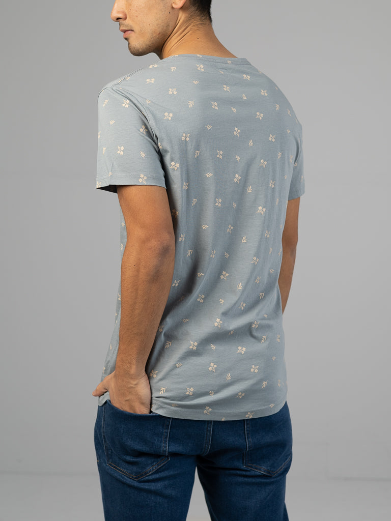 A person in a Heritage Printed Organic Cotton Tee by ID Menswear, featuring a light blue with small floral pattern and a relaxed fit, is seen from behind with hands in blue jeans against a plain gray backdrop.