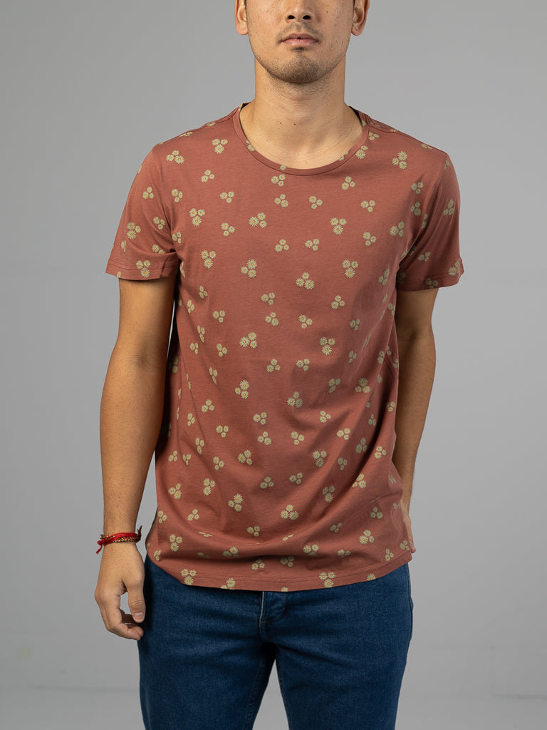 A person wearing the Heritage Printed Organic Cotton Tee by ID Menswear stands against a plain gray background. The rust-colored tee features a small floral pattern and is paired with blue jeans, with the persons hand resting on their hip.