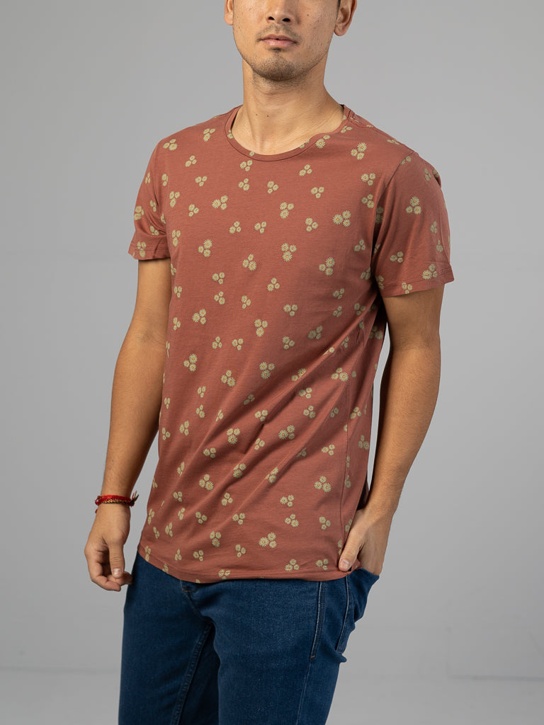 Someone wearing an ID Menswear Heritage Printed Organic Cotton Tee with a small floral pattern stands against a neutral background. Theyre also in blue jeans and have a red bracelet on their left wrist, with their head partially out of view.