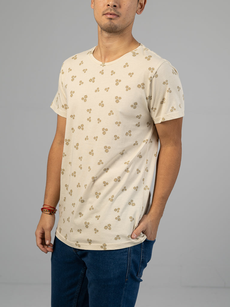 A man wears the ID Menswear Heritage Printed Organic Cotton Tee, a light beige T-shirt with a small floral pattern, paired with blue jeans. He has a bracelet on his right wrist and stands against a plain gray background, his face partially out of view.