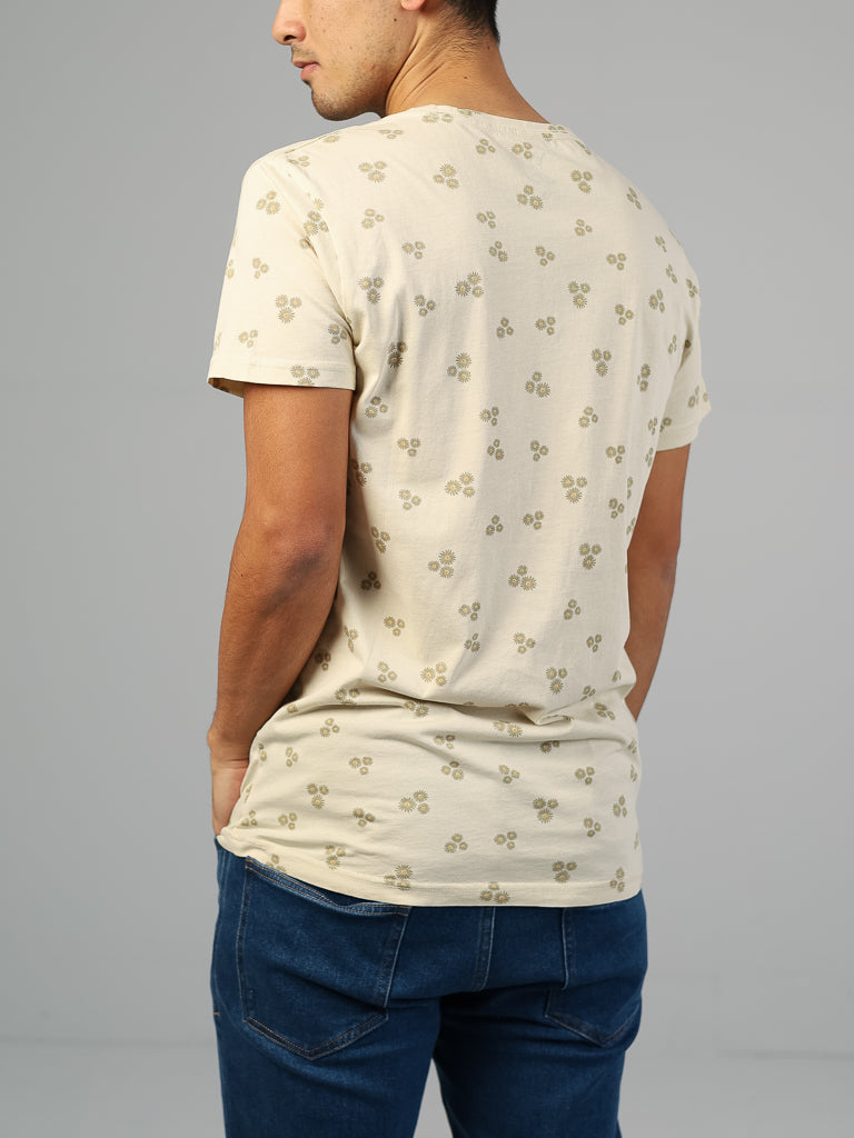 A person, back to the camera, wears the light cream Heritage Printed Organic Cotton Tee by ID Menswear featuring a small floral pattern, paired with dark blue jeans against a plain gray backdrop.