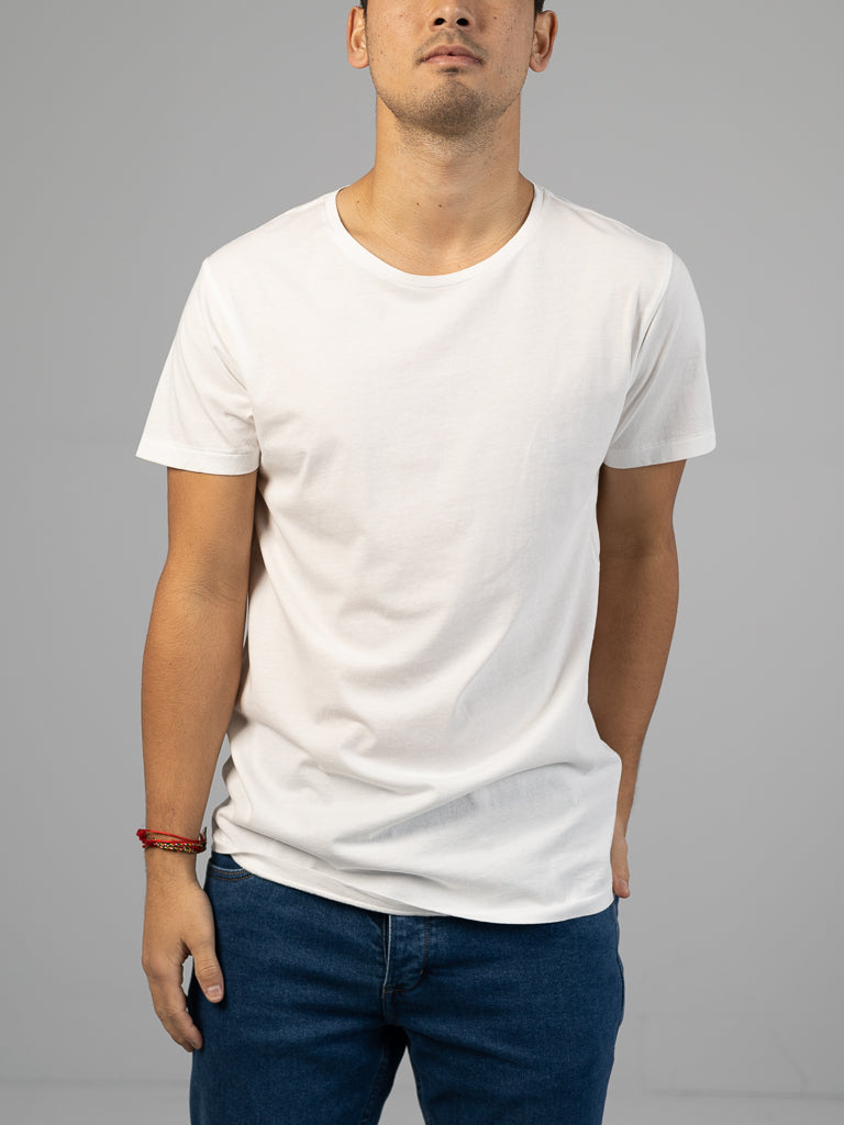 A person in a classic fit Legacy Organic Cotton Tee by ID Menswear and dark jeans stands against a neutral background. Their relaxed arms feature a red bracelet on the right wrist, subtly adding vintage flair to the serene setting.