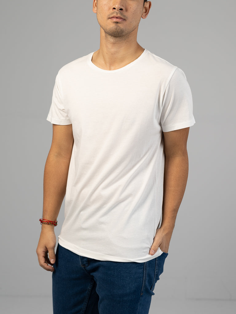 A person in an ID Menswear Legacy Organic Cotton Tee and blue jeans stands with one hand in their pocket against a gray background, accessorized with a red beaded bracelet on their left wrist, exuding a subtle vintage vibe.