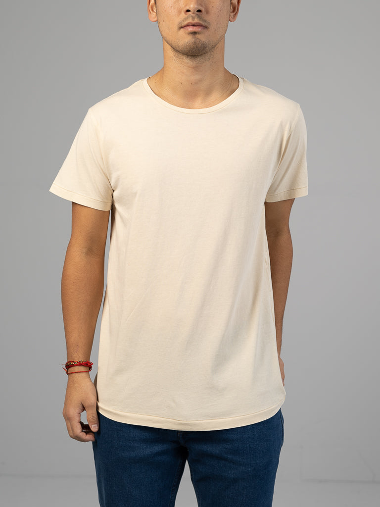 Someone wearing a Legacy Organic Cotton Tee by ID Menswear, classic fit in cream, paired with blue jeans stands against a neutral gray backdrop. Their face is hidden, but a red bracelet on their left wrist adds vibrant color to the vintage ensemble.