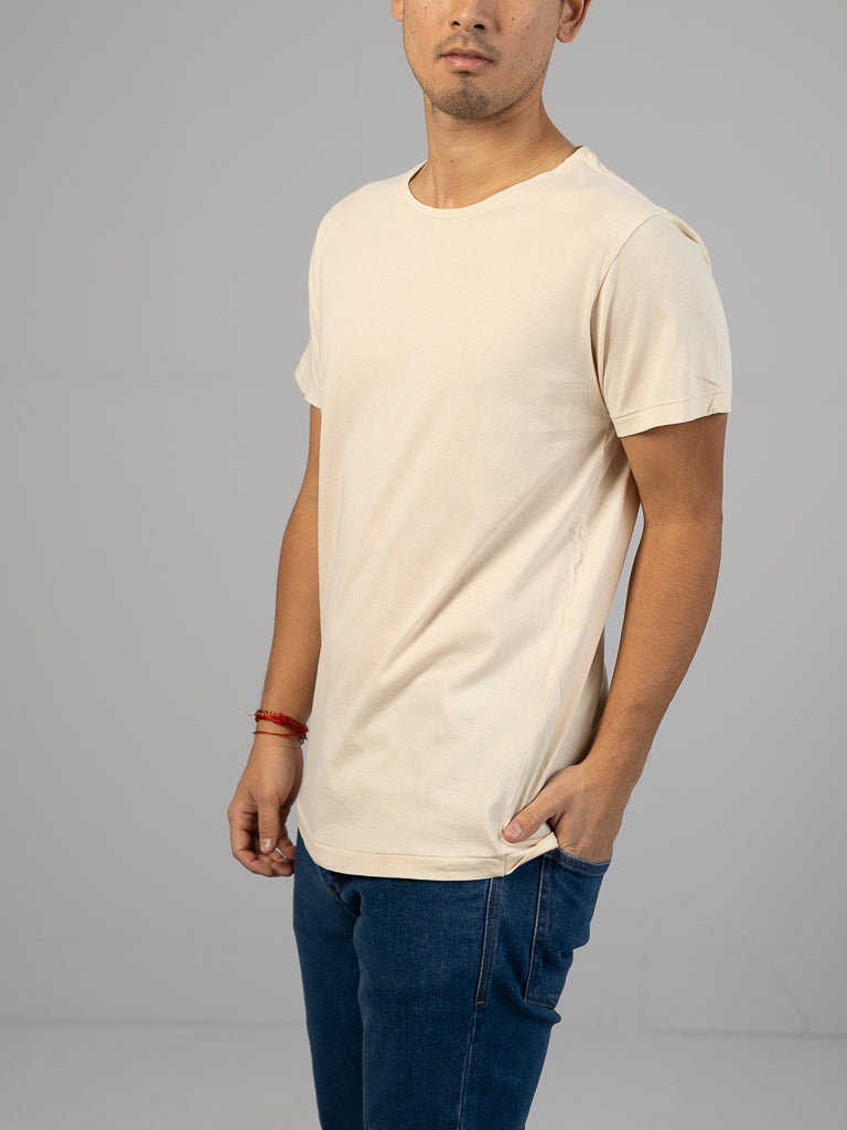 A person wears the Legacy Organic Cotton Tee by ID Menswear in beige, paired with blue jeans. They stand slightly sideways, left wrist adorned with a red bracelet and right hand in pocket. The classic fit enhances the vintage vibe against a plain gray background.