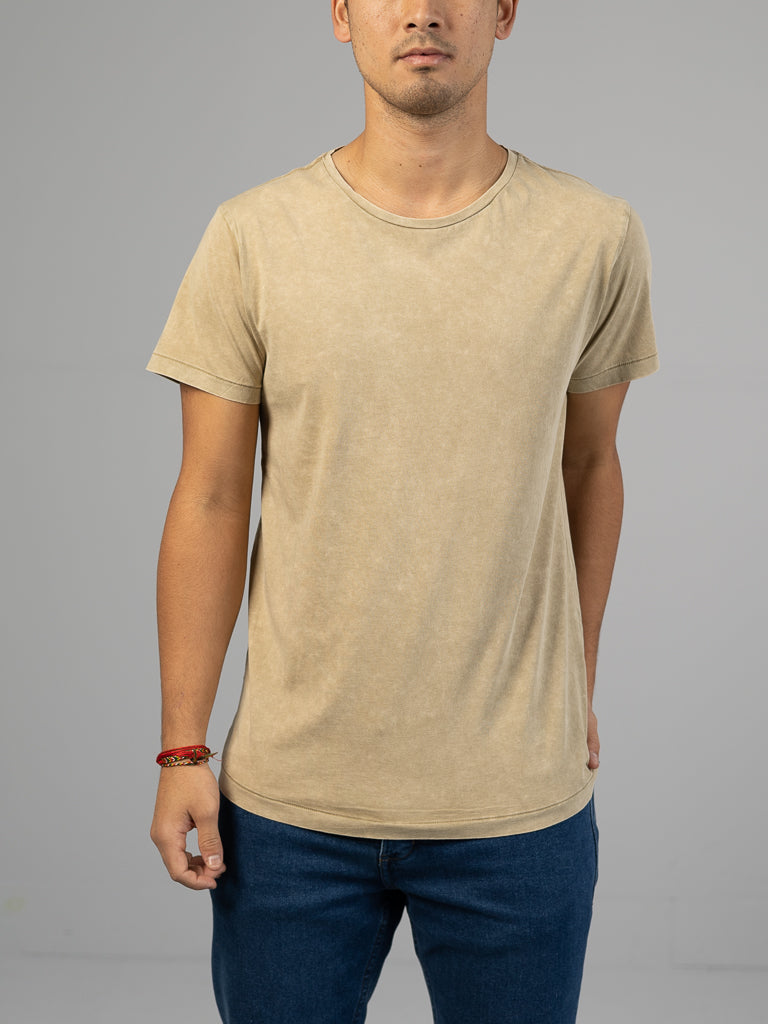Against a neutral gray backdrop, a person dons the ID Menswear Legacy Organic Cotton Tee in classic fit beige with blue jeans. Their left arm rests by their side, while a red bracelet adorns their right wrist.