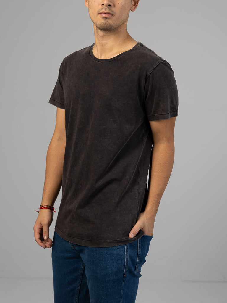 A person wearing the ID Menswear Legacy Organic Cotton Tee, a plain dark-colored shirt with a vintage look and classic fit, stands against a neutral background. They wear blue jeans and a red bracelet on their wrist. Their face is not visible as one hand rests in their pocket.