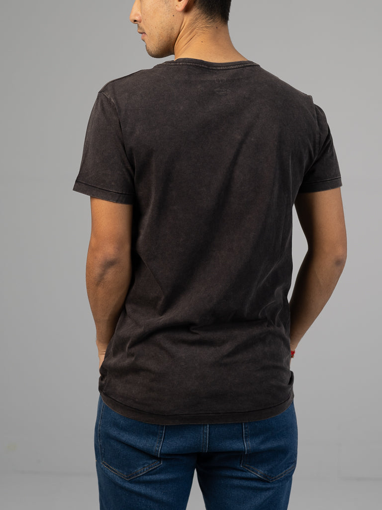 A person stands with hands in pockets against a gray background, wearing ID Menswears Legacy Organic Cotton Tee in dark gray and blue jeans, exuding a vintage look.