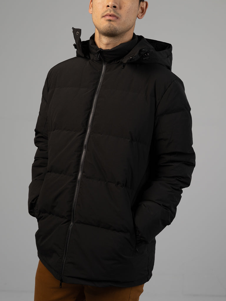 A person wears the ID Menswear Arctic Black Puffer Down Jacket with hands in pockets, paired with brown pants, standing against a plain gray background. Their face is partly visible.