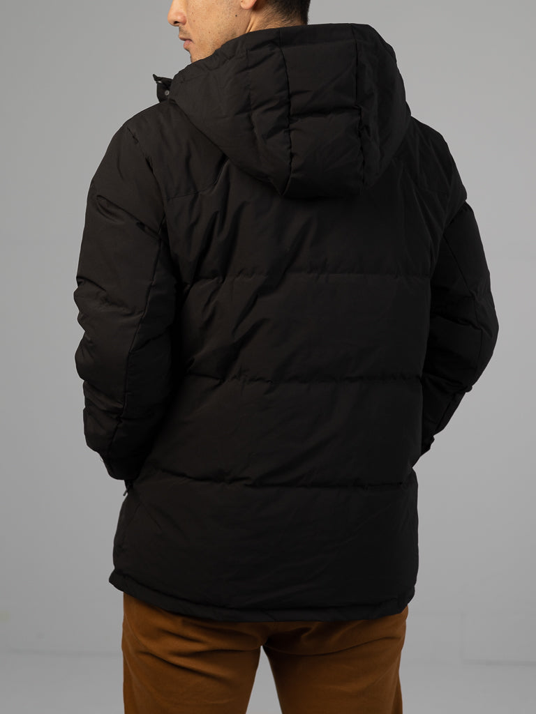 A person is seen from the back wearing the Arctic Black Puffer Down Jacket by ID Menswear with a hood, paired with brown pants. The background is plain and light-colored.