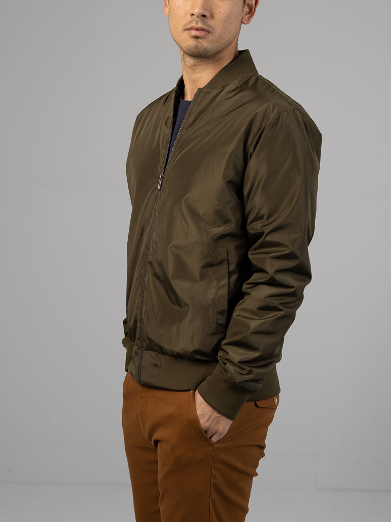 An individual in an olive-green Catskill Bomber Jacket from ID Menswear, zipped up with hands in pockets, stands against a neutral background in brown pants. The lightweight and water-resistant jacket obscures the face.
