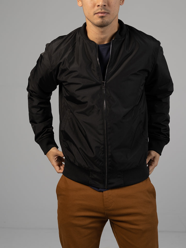 Someone in a black Catskill Bomber Jacket by ID Menswear and brown pants stands with hands in pockets against a simple, light gray background. The top of their head is slightly cropped out of the frame.
