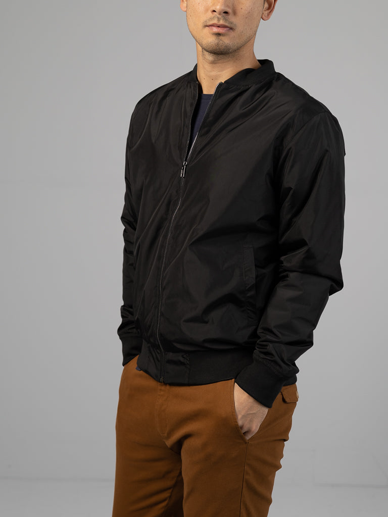 Wearing ID Menswears Catskill Bomber Jacket in black and brown pants, a person stands with hands in pockets against a neutral background, their face partially cropped out.