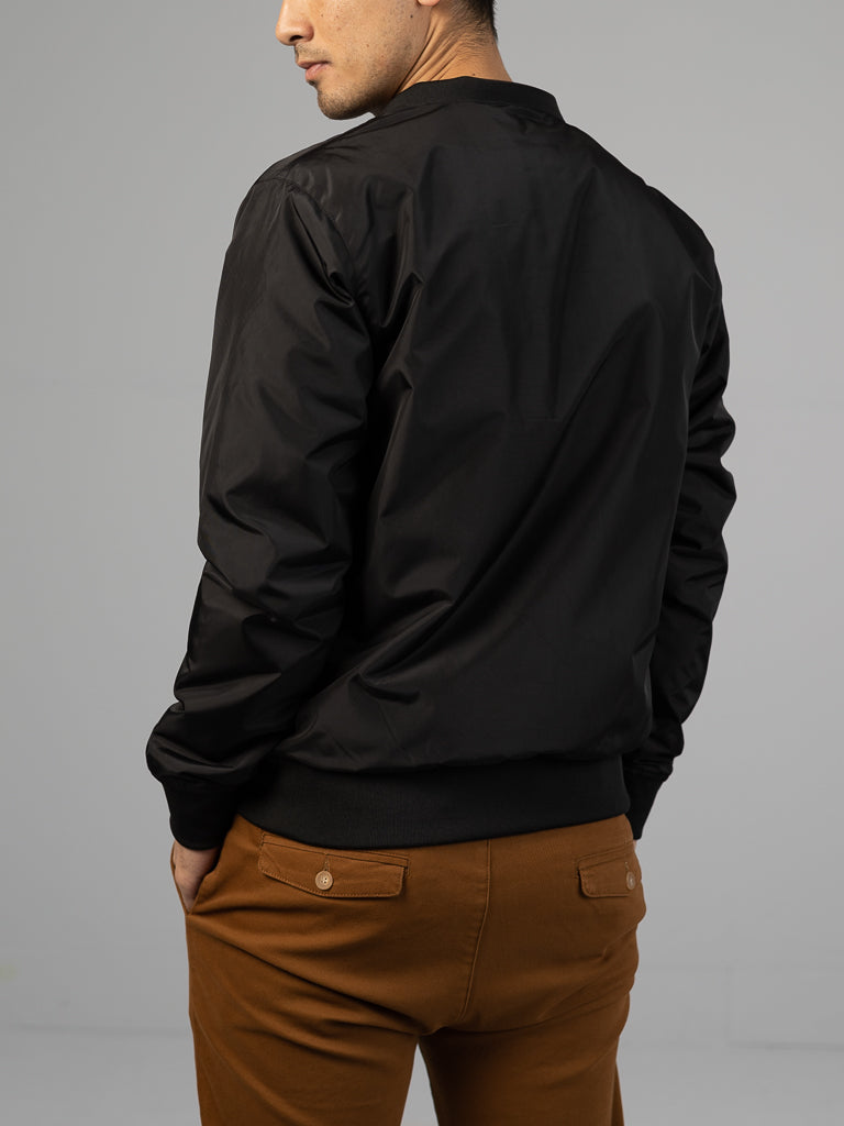 Wearing the Catskill Bomber Jacket, a lightweight black piece by ID Menswear, a person stands facing away in brown pants. They have short, dark hair against an uncluttered gray background.