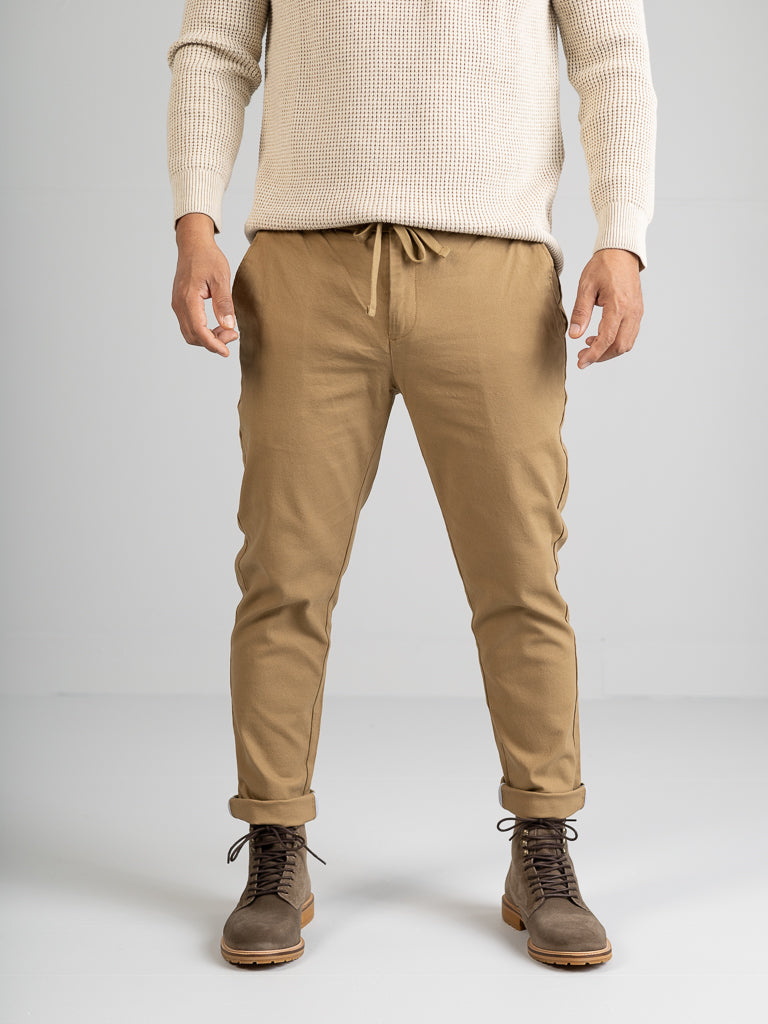 A person wearing a cream sweater and ID Menswears Kent Drawstring Canvas Pant in tan stands against a plain background. They also sport brown leather boots, with their hands relaxed by their sides.