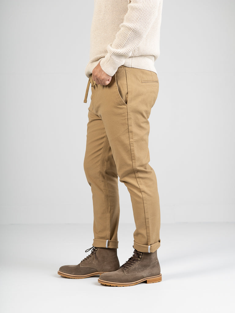 A person is wearing ID Menswears Kent Drawstring Canvas Pant with brown leather boots. The cream knit sweater completes the look, as they stand against a plain white backdrop with one hand in their pocket. Only the lower half of the outfit is visible.